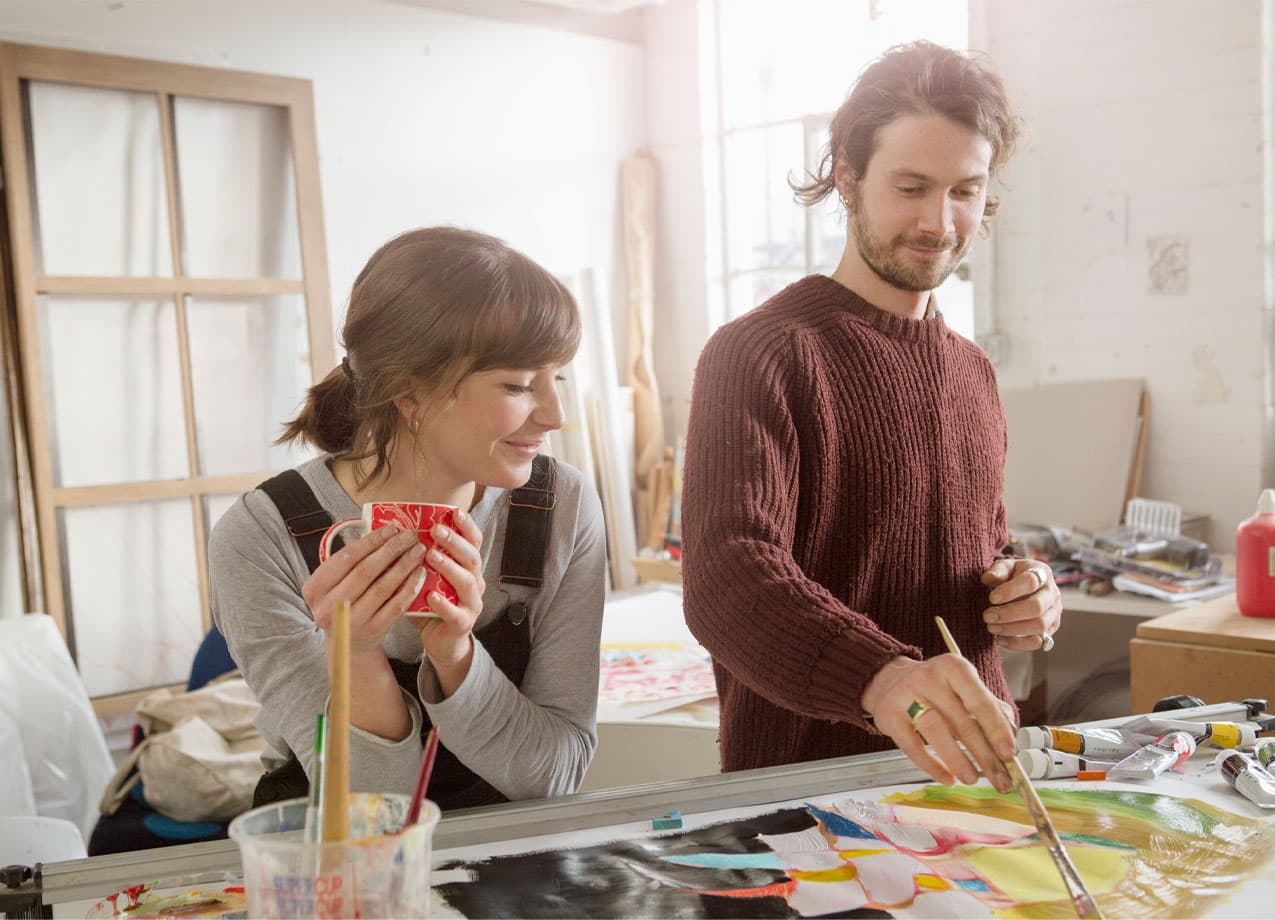 hobbies-for-couples: A man and a woman stand in an art studio at a table. The man is painting and the woman is leaning over the table holding a mug watching.