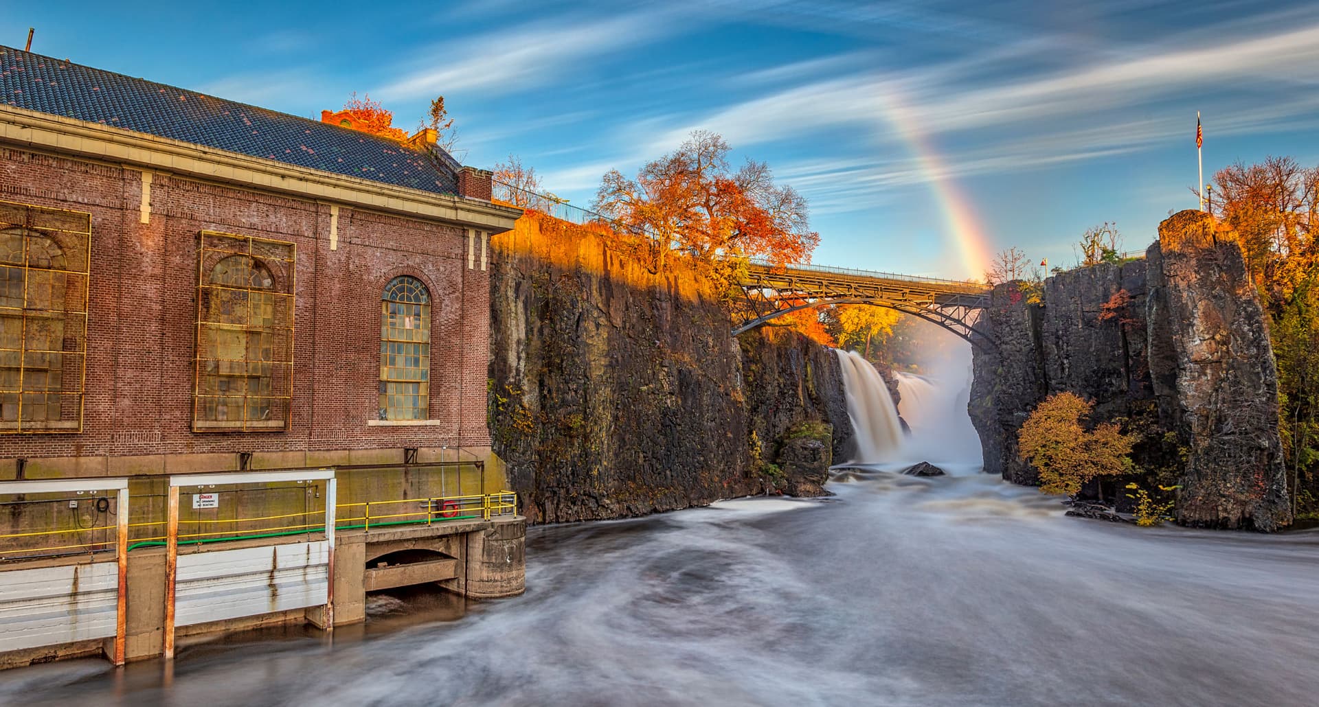 Hidden Gems NJ - A large older building sits on the edge of a river contained by two stone cliffs. There's a bridge connecting the two stone cliffs and waterfalls just behind it. The sky is a bright blue with clouds and a rainbow.