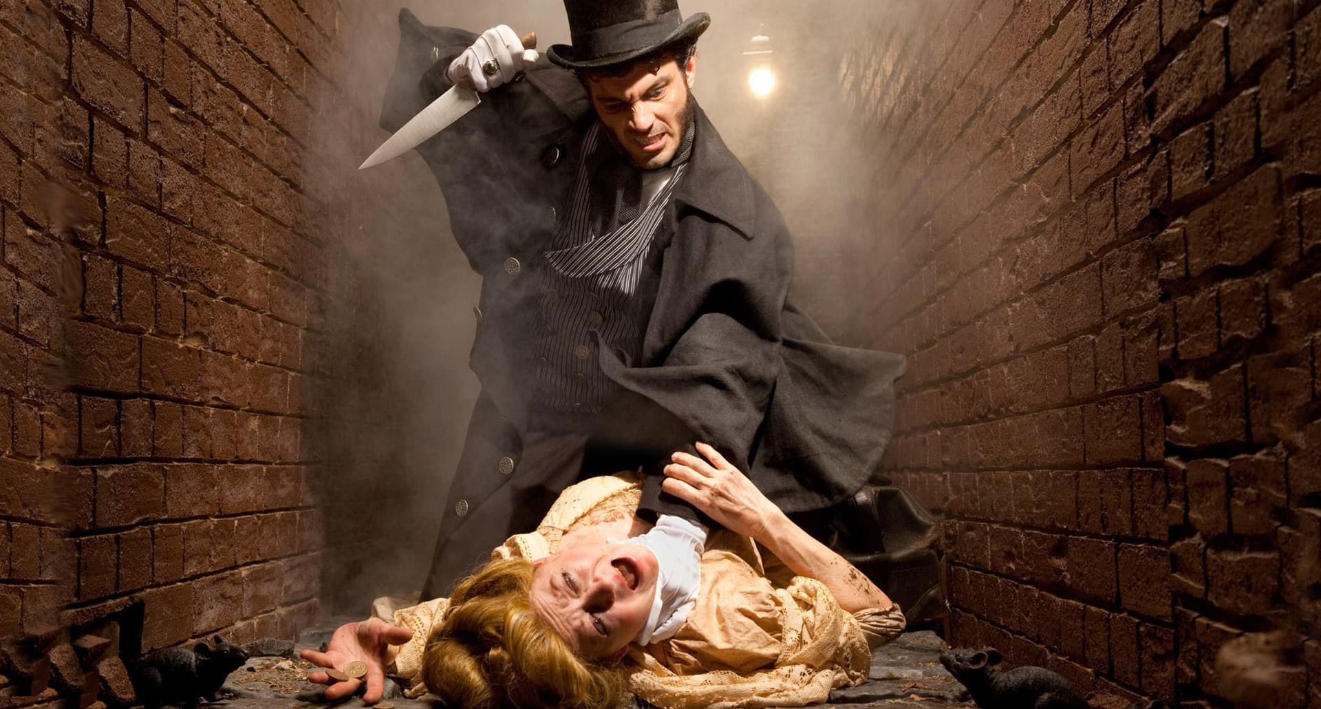 NY Haunted Houses - A man dressed in 1800s style clothes with a coattails and a top hat climbs over a woman with a dagger in his hand as he attempts to murder her. They are in a brick lined alley way. The woman is dressed in an old fashioned yellow dress and has her hair up in a loss updo.