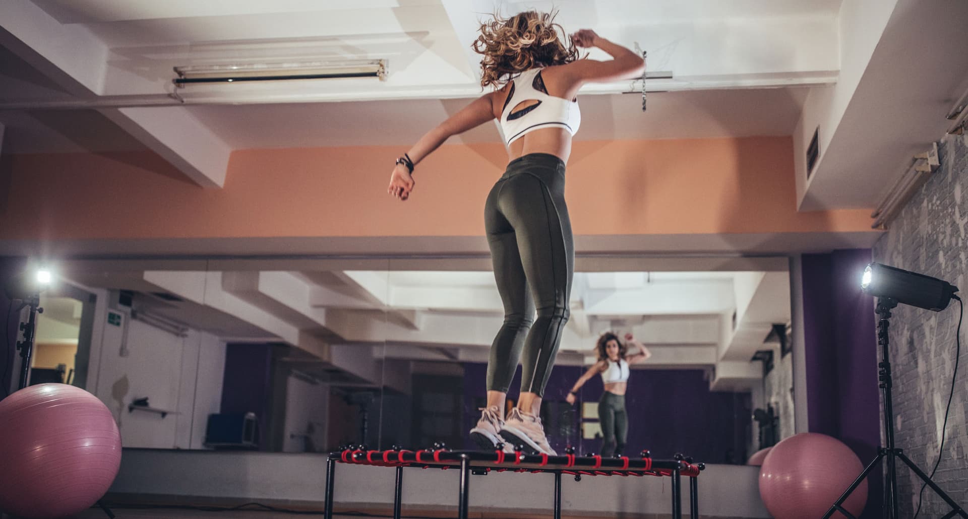 Trampolining San Francisco - A white woman dressed in workout gear jumps on a mini trampoline in a workout studio. You can see their reflection in a mirror at the front of the studio.
