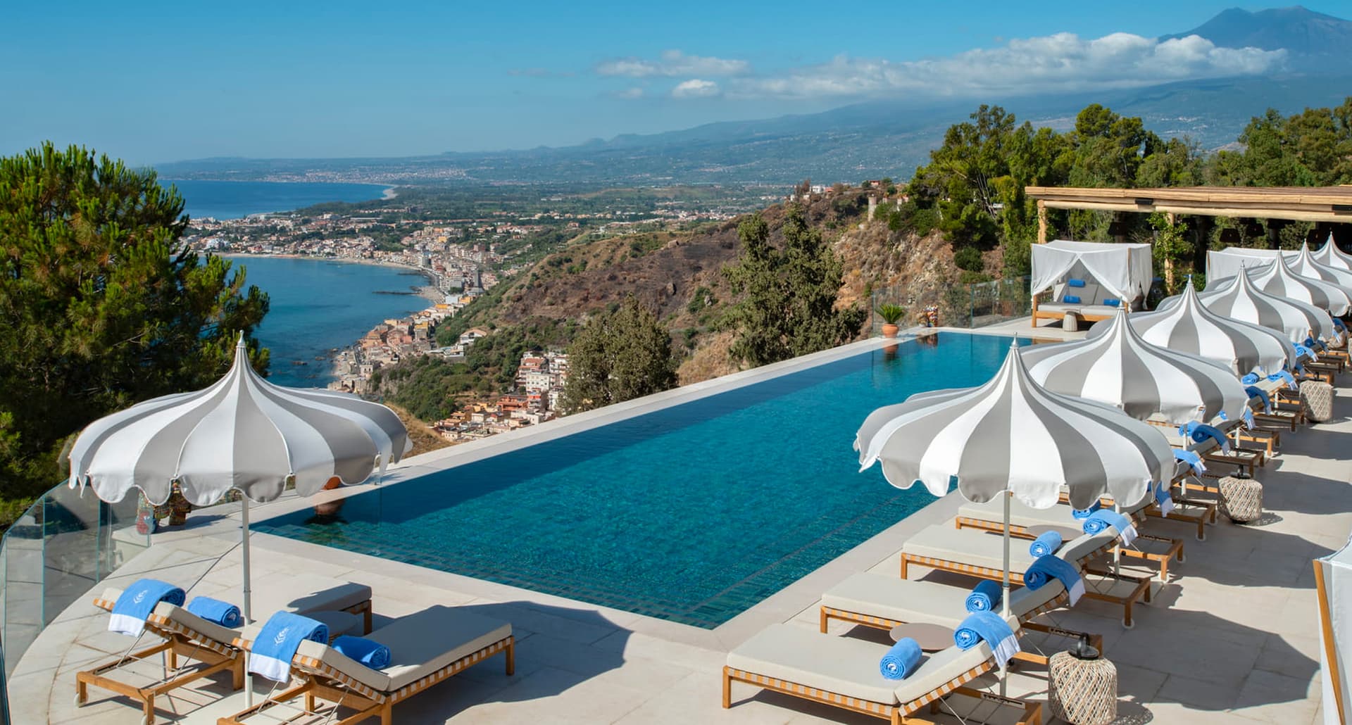 Places Like Sicily - A photograph of a very blue, long rectangular pool that overlooks a number of trees, the ocean, and houses. The pool is surrounded by lounge chairs with ornate striped umbrellas.