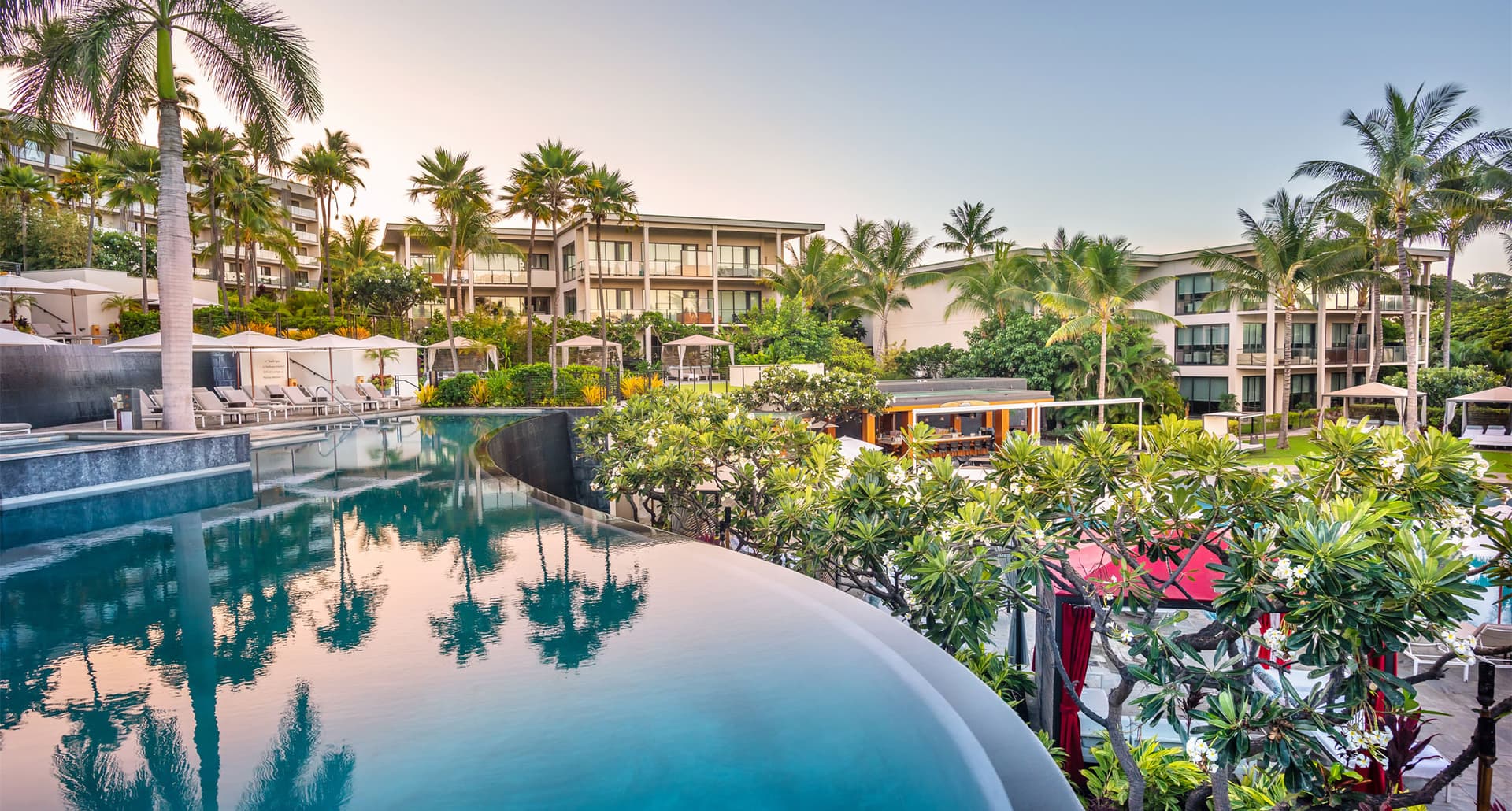 Best Hotels in Wailea Hawaii - A photograph of the Andaz Maui hotel pool. There's a reflection of palm trees in the pool. It's surrounded by plants, flowers and other vegetation. There are buildings from the property in the distance.