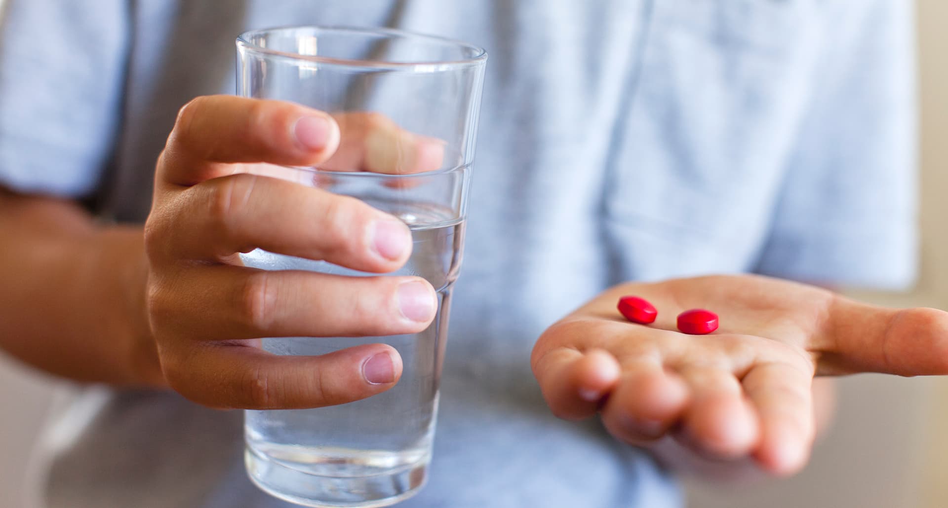 should i give my child adhd medication: close up of boy holding a glass of water in one hand and medication pills in the other hand.