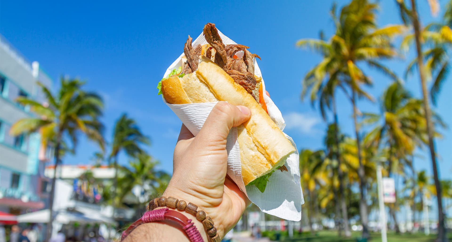 Best Food in Miami - A person holds up a big Cuban sandwich in front of a street lined with palm trees. The sky is bright.