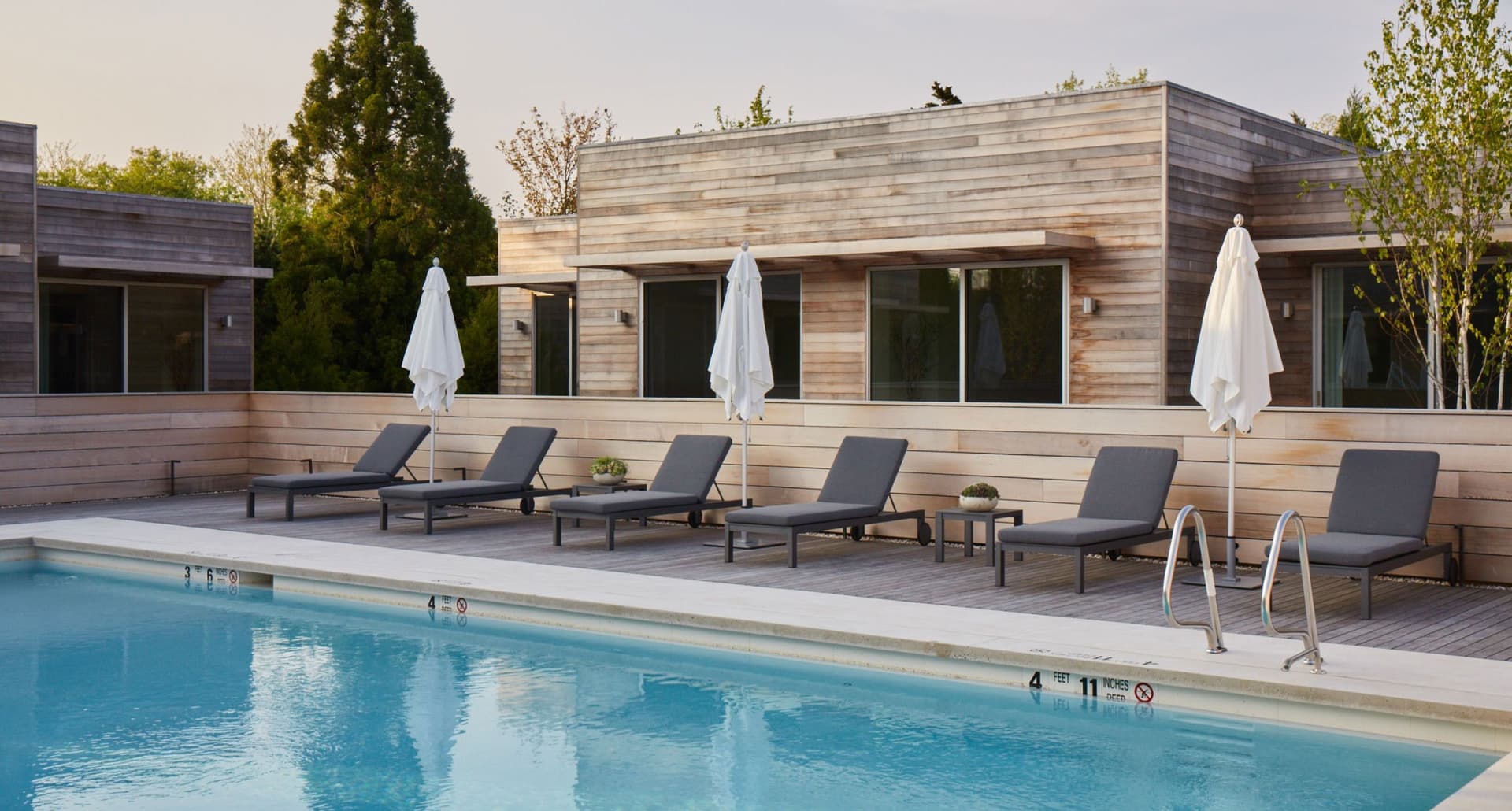 best-spas-in-the-hamptons: A photograph of an outdoor pool at a spa. There are gray lounge chairs surrounding the pool and a wooden building behind the pool.