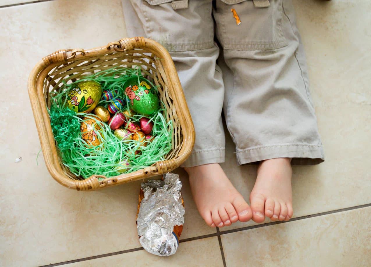 best-premade-easter-baskets: A photo of a young child's feet next to an Easter basket of chocolate eggs on fake grass stuffing. A large tinfoil wrapper sits next to the boy and the basket.