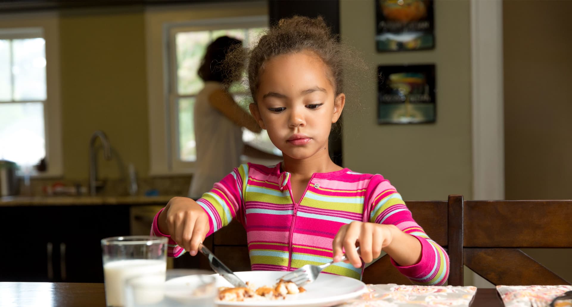 how-to-teach-kids-manners: A young girl of color sits at a dinner table and uses a pair of utensils to cut something on her plate. There is a glass of milk on the table. She wears a pink and green striped long sleeve shirt. In the background, you can see an adult in the kitchen.