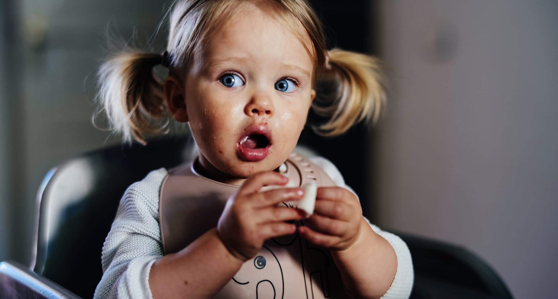 A toddler baby girl eats food with a look of shock on her face.
