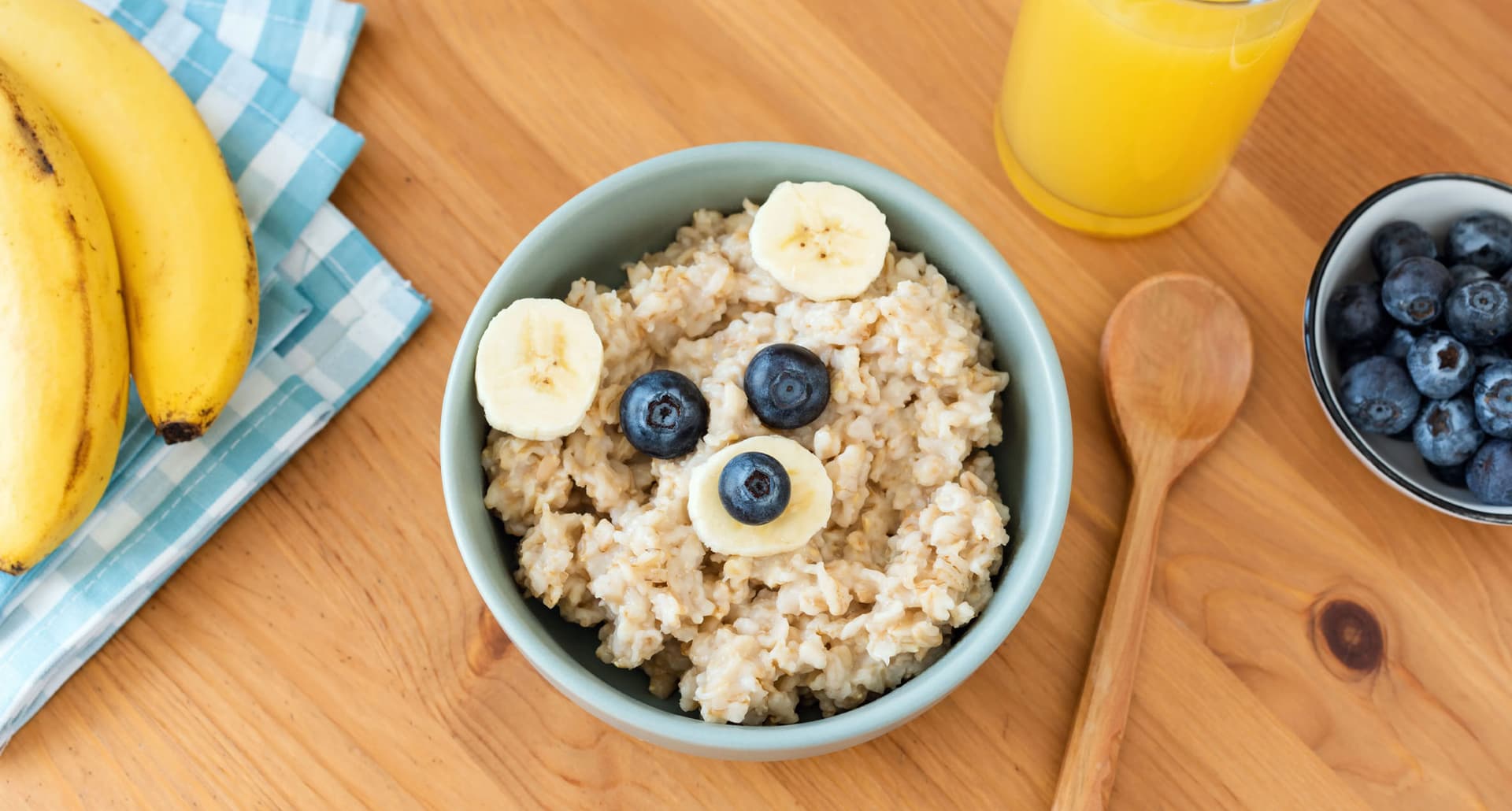 high-fiber-foods-for-kids: A photo of a bowl of oatmeal topped with blueberries and bananas. Next to it is a glass of OJ, a bowl of blueberries, a bunch of bananas, a wooden spoon and a plaid napkin.