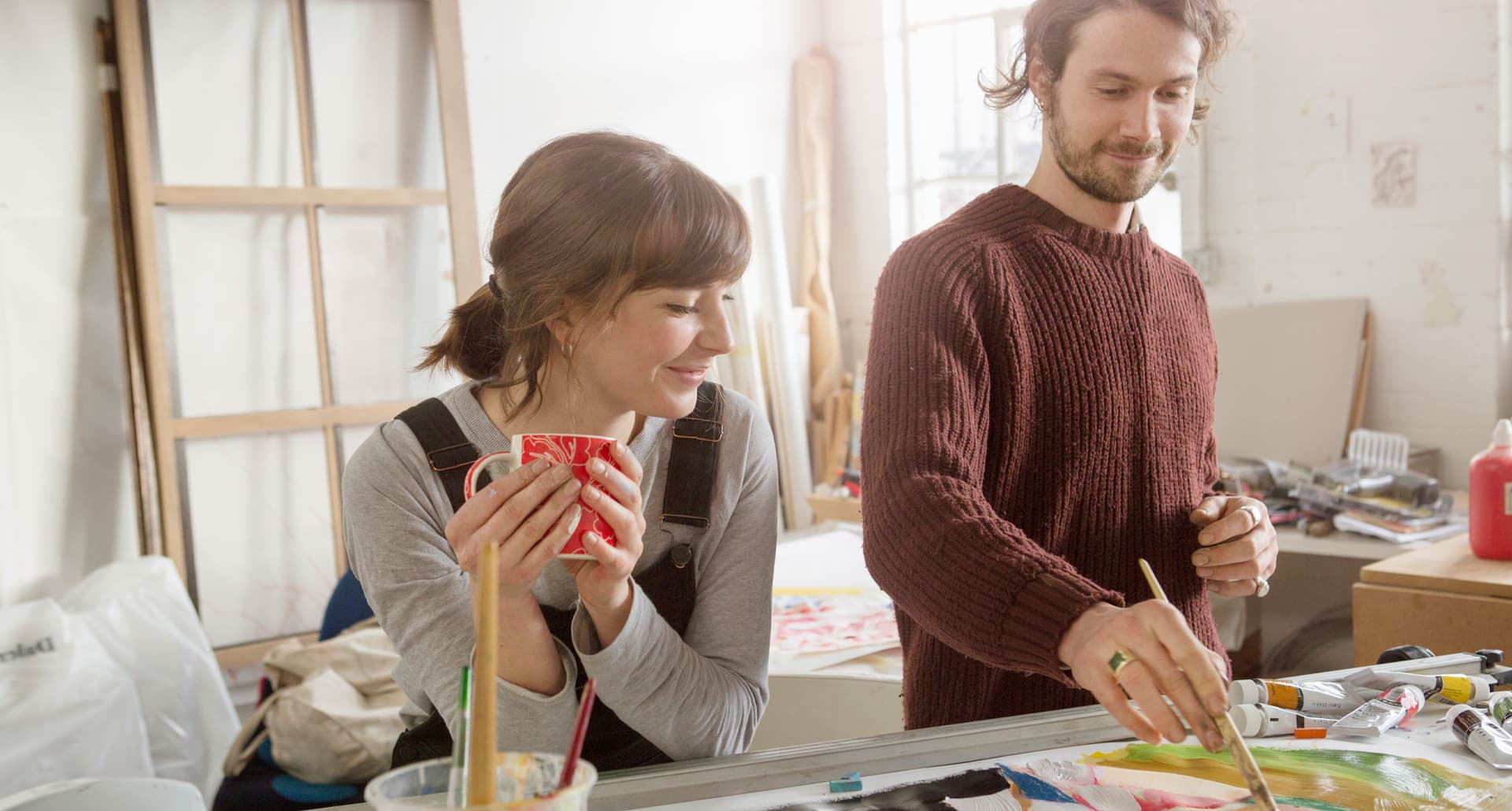 hobbies-for-couples: A man and a woman stand in an art studio at a table. The man is painting and the woman is leaning over the table holding a mug watching.