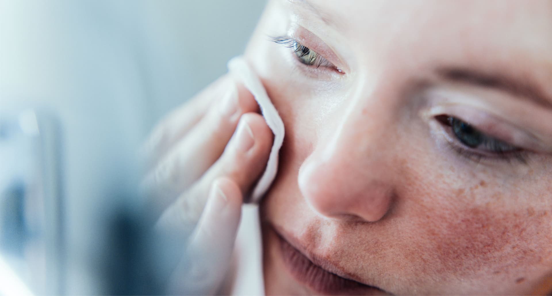 10 Products for Sensitive Skin - A close up shot of a pale white woman with freckles wiping her face with a cotton round. She is looking in a mirror.