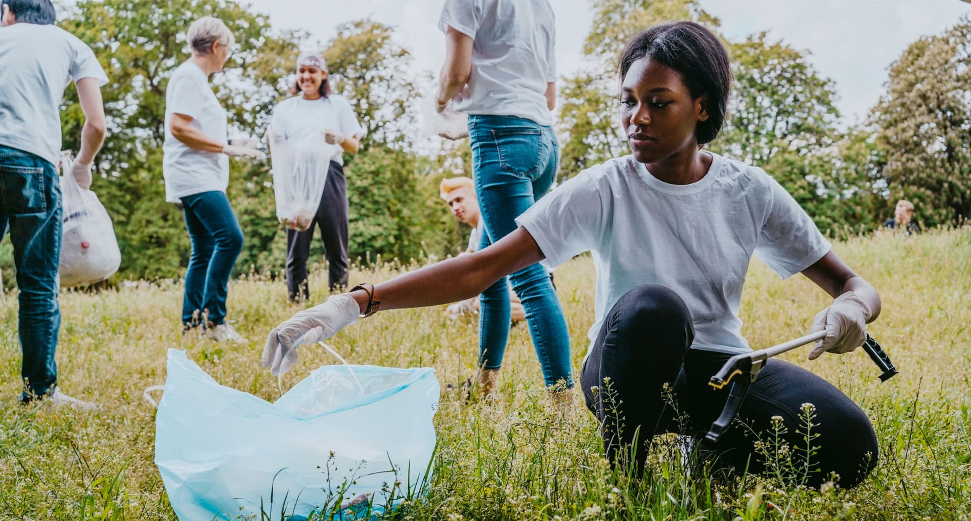 LA Charities - A young Black woman in a white shirt and rubber gloves picks up garbage in a grassy field. There is a light blue trash bag next to her and a group of people behind her also picking up garbage.