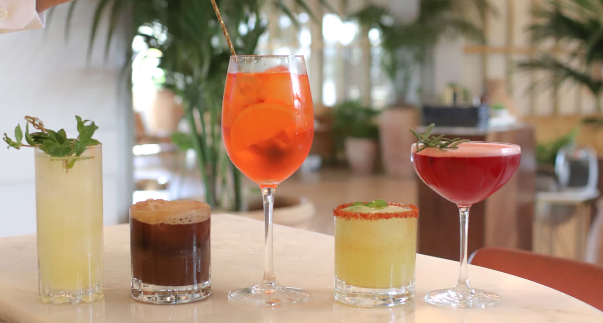 Non Alcoholic Drinks LA - A row of five brightly colored non alcoholic drinks are lined up on a countertop. They are each in differently shaped glasses of varying heights. From left to right: A light yellow drink in a cylindrical glass, a brown drink in a whiskey glass, an orange drink in a wine glass, a lime green drink in a whiskey glass, and a bright red drink in a coupe glass.