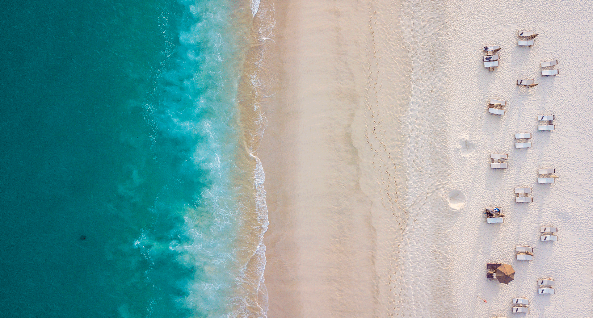 Things to Do in Los Cabos - An overhead shot of a white sand beach where it means with bright blue green water. Some people and chairs dot the sand.