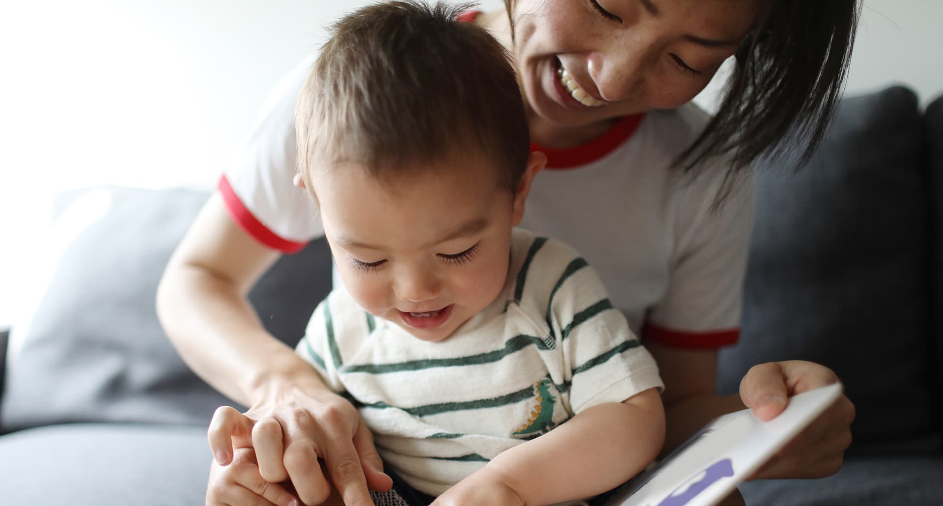 toddler-speech-milestones-ask-a-pediatrician: A young mother and her son sit together reading a book. They seem to be of Asian descent. She wears a white shirt with red ringer sleeves and the son wears a white shirt with green stripes.