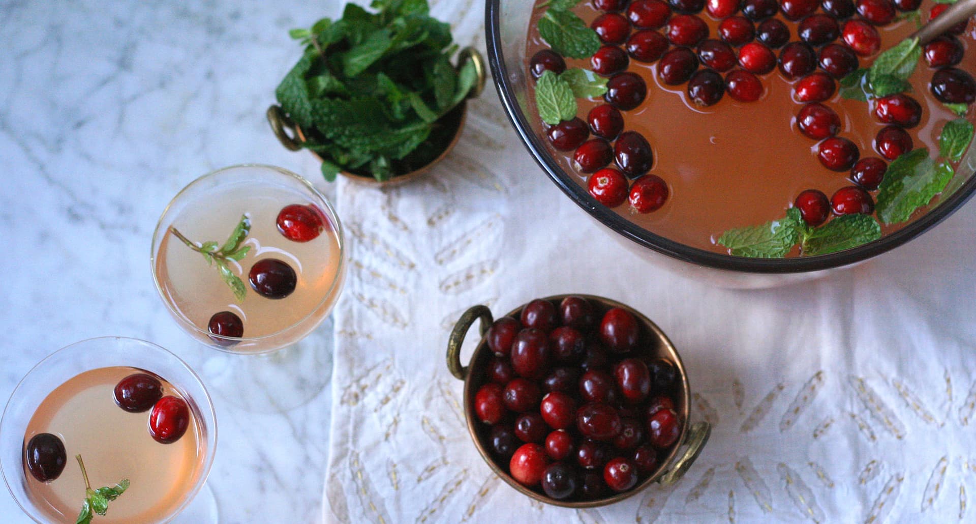 holiday punch bowl cocktails: cranberry mint cocktail in a bowl with two glasses and a bowl of cranberries