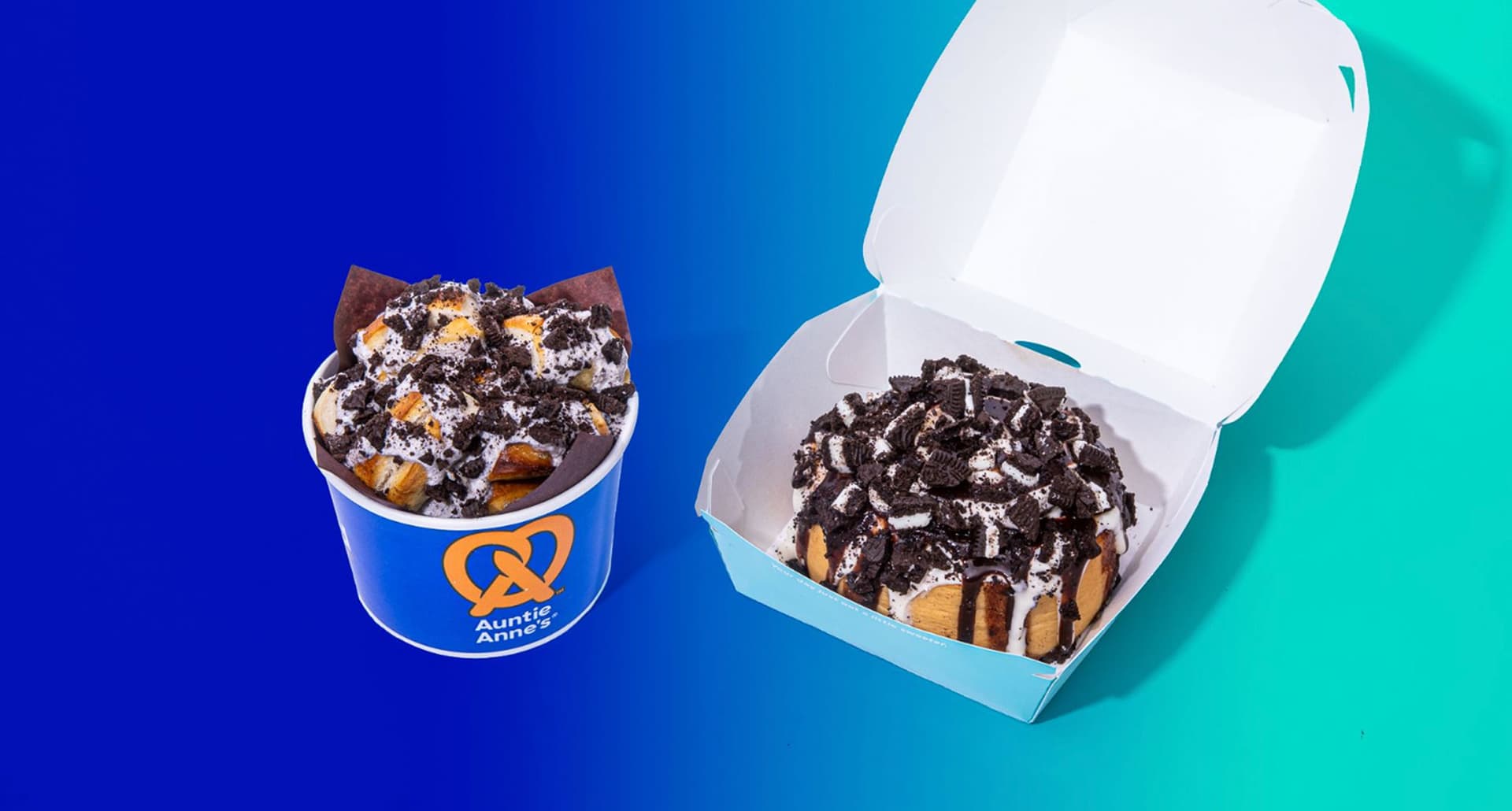 auntie-annies-cinnabon-oreo-topped-nuggets-and-buns: A photograph of Auntie Anne's pretzels and a Cinnabon cinnamon bun topped with OREO glaze and crumble. They are in front of a blue and sea foam green ombre background.